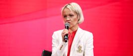 Kobieta w białej marynarce na czerwonym tle przemawia przez mikrofon
