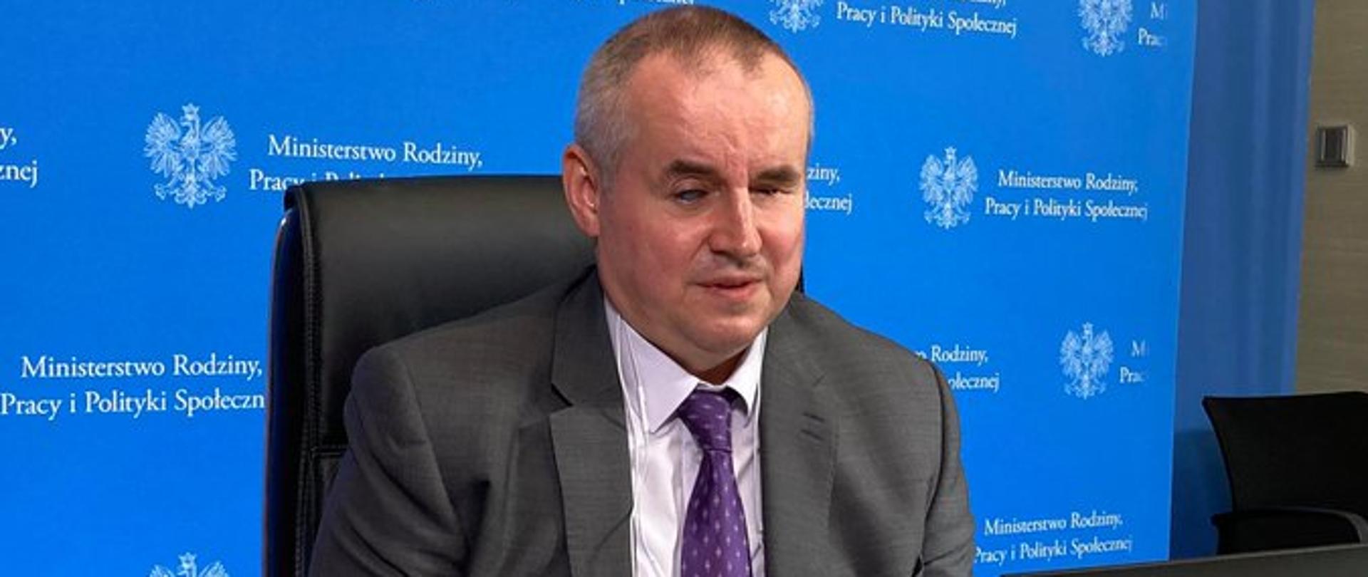 Wiceminister Paweł Wdówik podczas wideokonferencji. Przed nim na stoliku laptop. Za plecami baner z logiem Ministerstwa Rodziny, Pracy i Polityki Społecznej poprzedzony godłem.