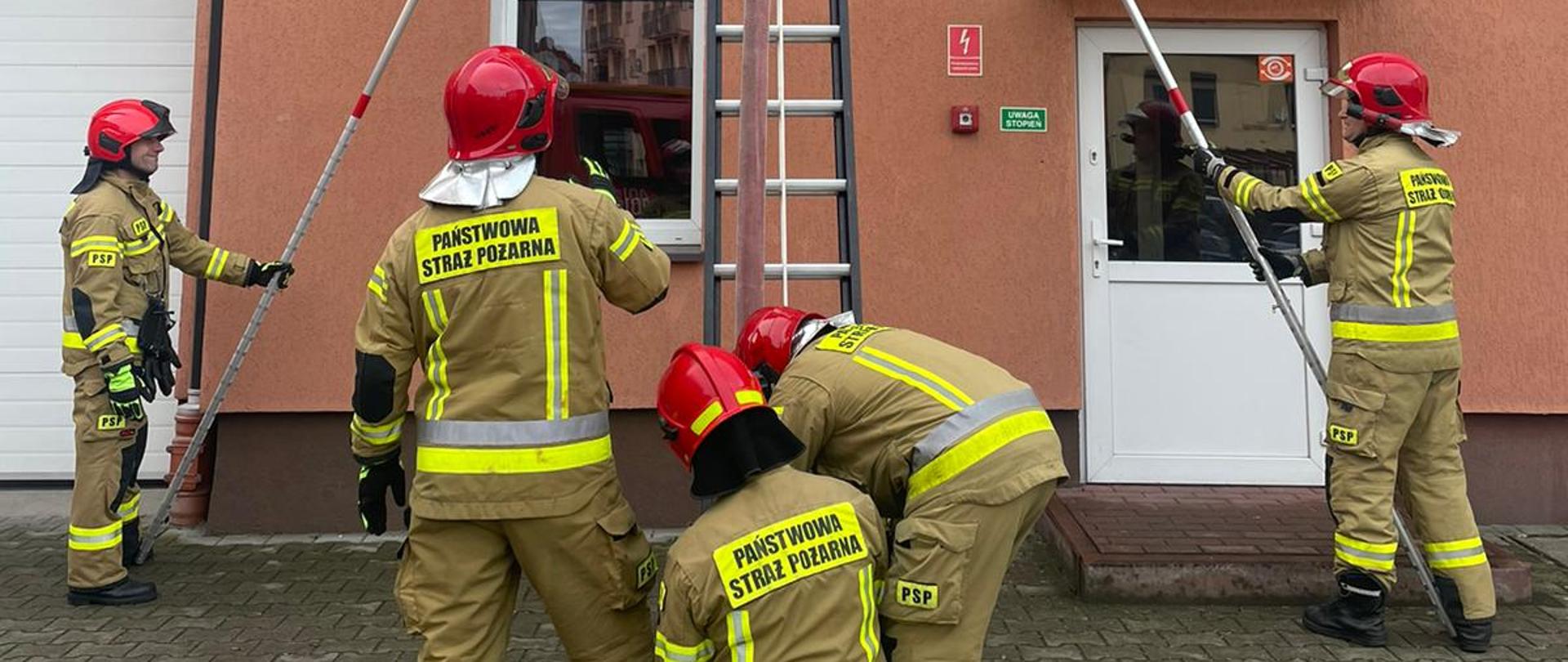Zdjęcie przedstawia strażaków ćwiczących na placu z drabinami przenośnymi.
W tle budynki.
