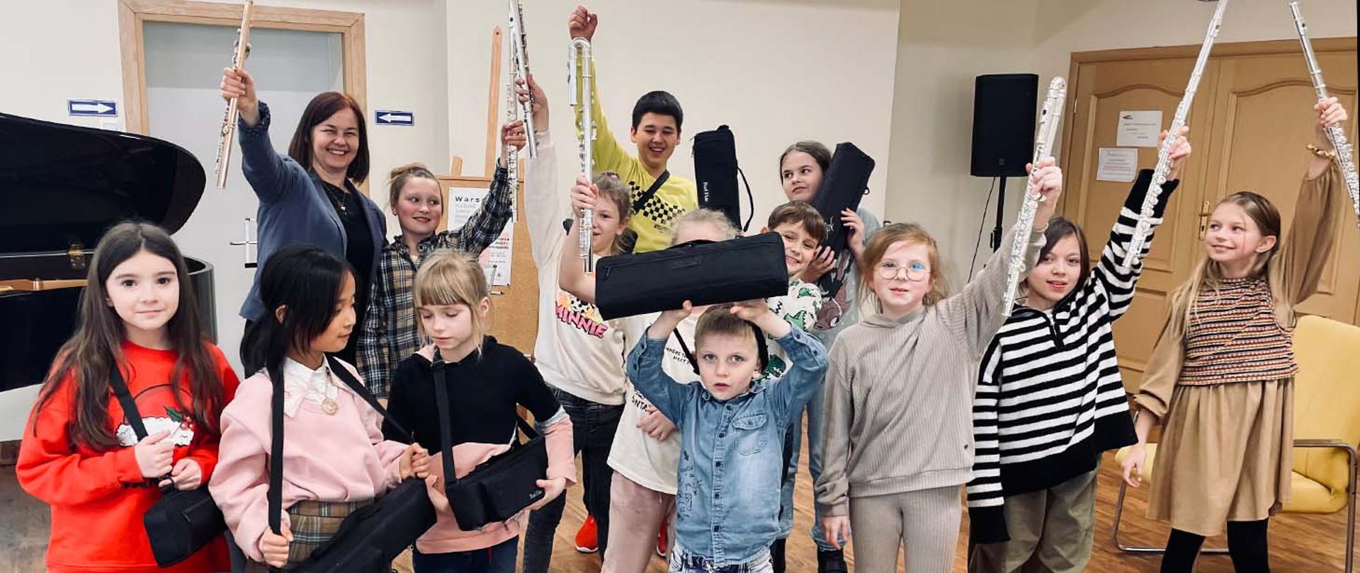 Uczestnicy warsztatów - 13 dzieci i nauczycielka stoją w auli, wszyscy w uniesionych do góry rękach trzymają flety.