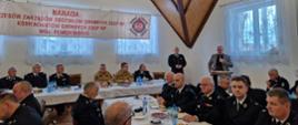 Strażacy ochotniczych straży pożarnych oraz Państwowej Straży Pożarnej siedzą za stołami mężczyzna stoi przed mównicą i przemawia trzymając mikrofon.