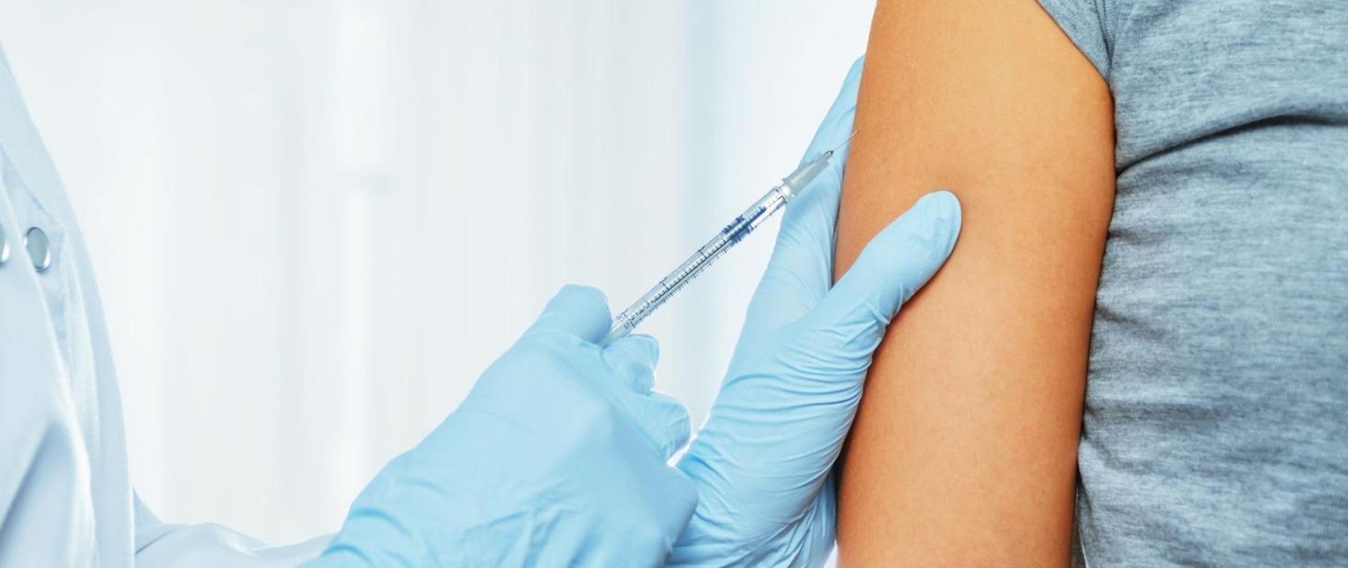 Na zdjęciu widoczne jest ramię pacjenta oraz dłonie lekarza. Medyk wykonuje szczepienie przeciw COVID-19 za pomocą trzymanej w ręce strzykawki.