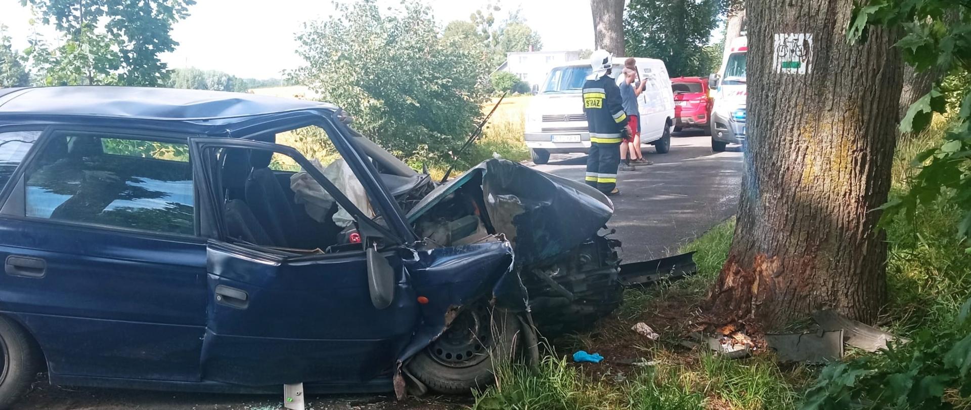 Wrak samochodu osobowego po uderzeniu w drzew, w tle strażak i osoby postronne