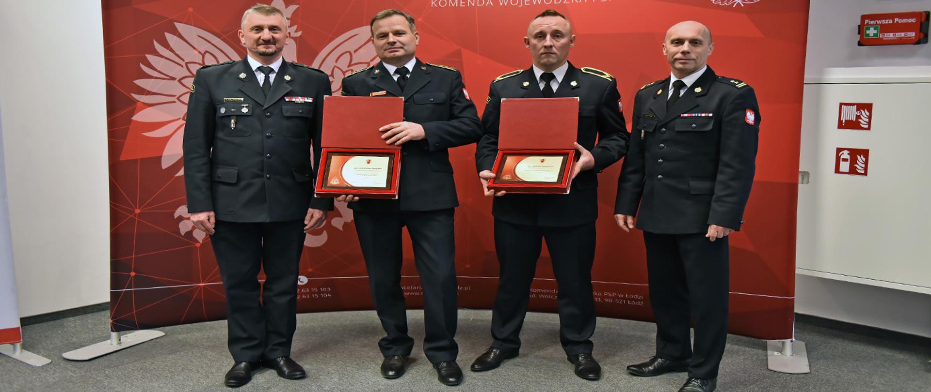 czterech funkcjonariuszy państwowej straży pożarnej ubranych w mundury wyjściowe pozuje do zdjęcia, dwóch trzyma w ręku pamiątkowe tabliczki z tytułem najlepszego dyżurnego stanowiska kierowania psp oraz najlepszego strażaka psp, po prawej stronie stoi komendant powiatowy brygadier Arkadiusz Makowski a po lewej zastępca komendanta wojewódzkiego starszy brygadier Paweł Malinowski, w tle widoczna ścianka barwy czerwonej z białymi napisami PAŃSTWOWA STRAŻ POŻARNA KOMENDA WOJEWÓDZKA PSP W ŁODZI z godłem państwowej straży pożarnej oraz symbolem orał białego