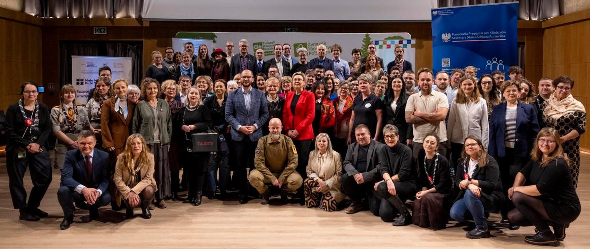 Konferencja regionalna programu Moc Małych Społeczności w Pabianicach. Szeroki kadr, zdjęcie grupowe. Uczestnicy wydarzenia pozują do wspólnego zdjęcia, prezentując otrzymane Certyfikaty Twórcy Odporności Społecznej. W środkowej części zdjęcia widoczni Sekretarz Stanu Adriana Porowska oraz Michał Braun, Dyrektor Narodowego Instytutu Wolności. 