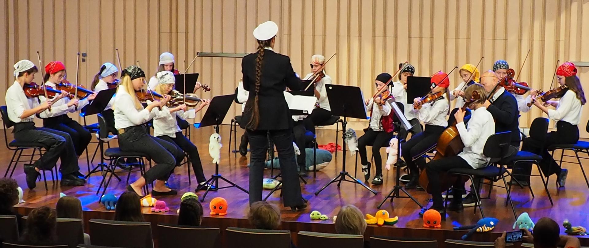 Młodzieżowa orkiestra smyczkowa z dyrygentem na scenie w sali koncertowej.