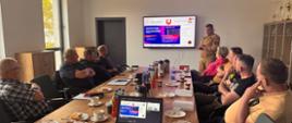 Na zdjęciu widać grupę osób siedzących przy stole konferencyjnym. Patrzą się oni na prezentacje z OC i OL, którą prezentuje Komendant Powiatowy PSP.