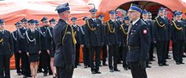Zdjęcie przedstawiające grupę strażaków w czarnych, galowych mundurach na placu. Na pierwszym planie dwóch oficerów stoi naprzeciwko siebie w postawie na baczność, z których jeden, po prawej stronie, melduje. W tle widać rzędy strażaków oraz pomarańczowe namioty.