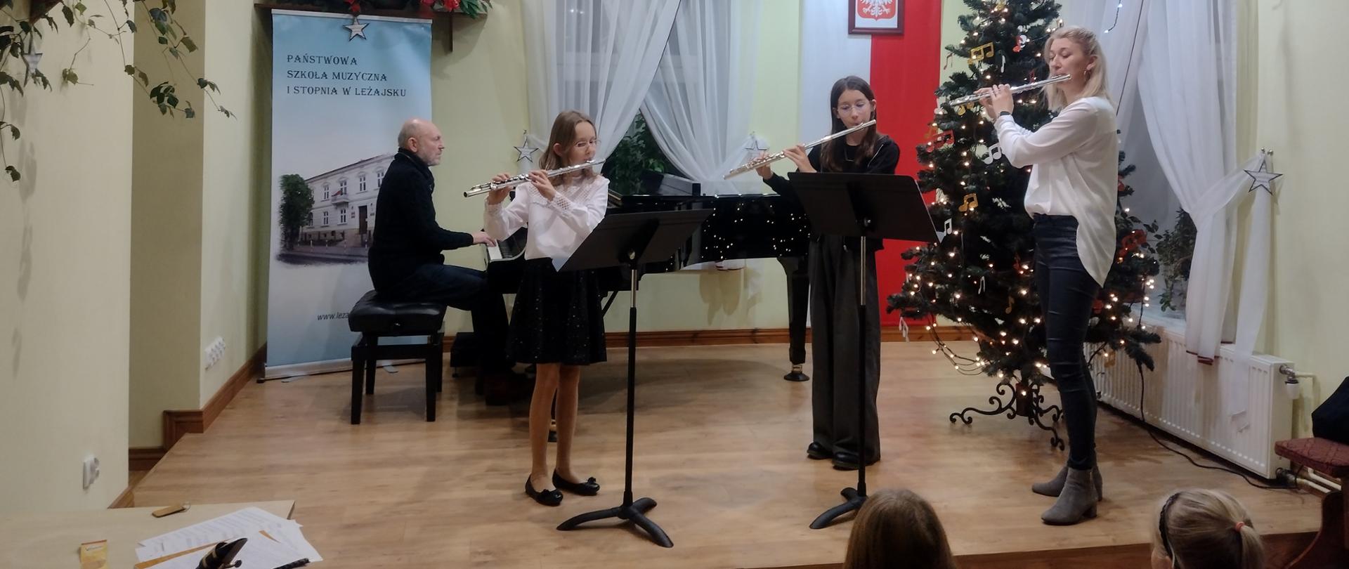Trio fletowe na scenie w trakcie gry. Z tył występujących czarny fortepian, przy nim akompaniator. Po prawej stronie zdjęcia za jedną z grających flecistek udekorowana choinka. Zdjęcie utrzymane w jasnych kolorach.