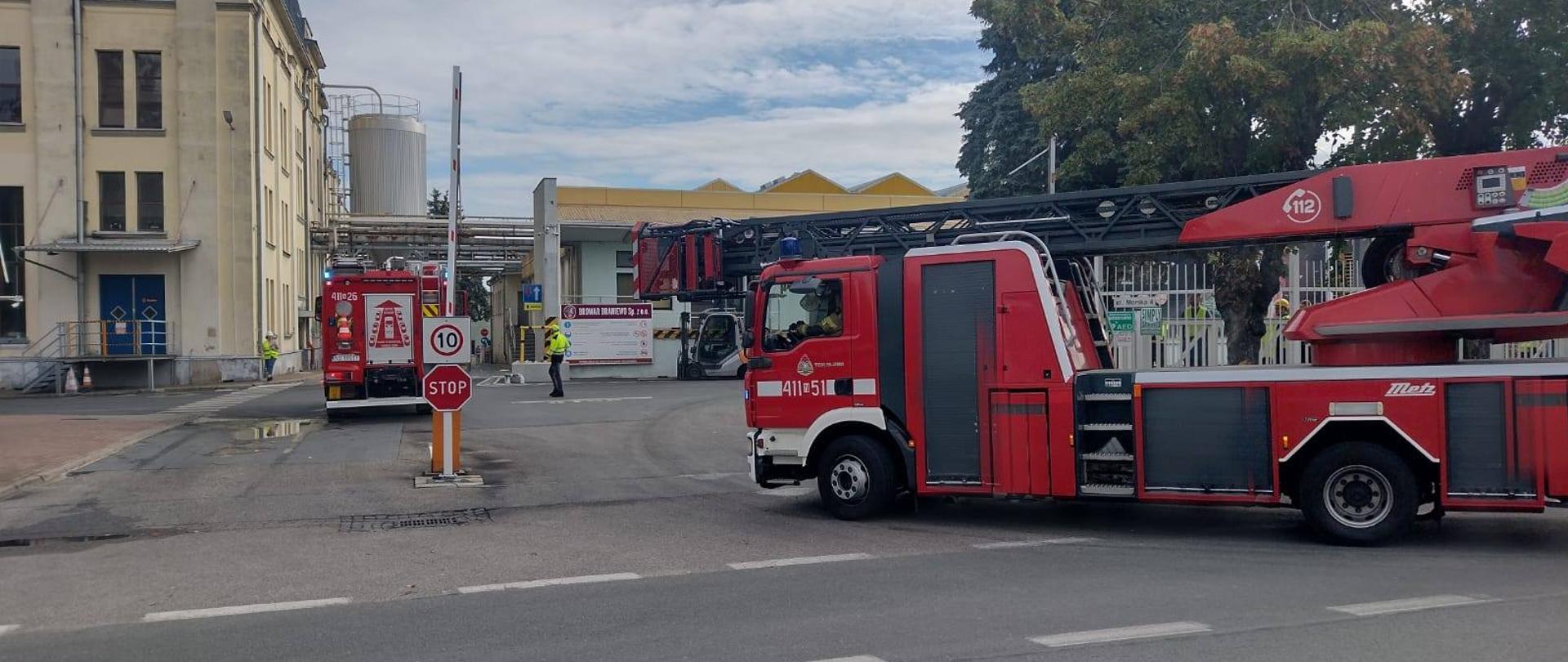 Zdjęcie przedstawia akcję straży pożarnej – na terenie zakładu przemysłowego. Na pierwszym planie widoczne są dwa wozy strażackie, w tym jeden z drabiną mechaniczną, a w tle kolejny samochód gaśniczy. Strażacy zabezpieczają teren i prowadzą działania. Na miejscu obecni są także pracownicy zakładu i osoba kierująca ruchem przy wjeździe.