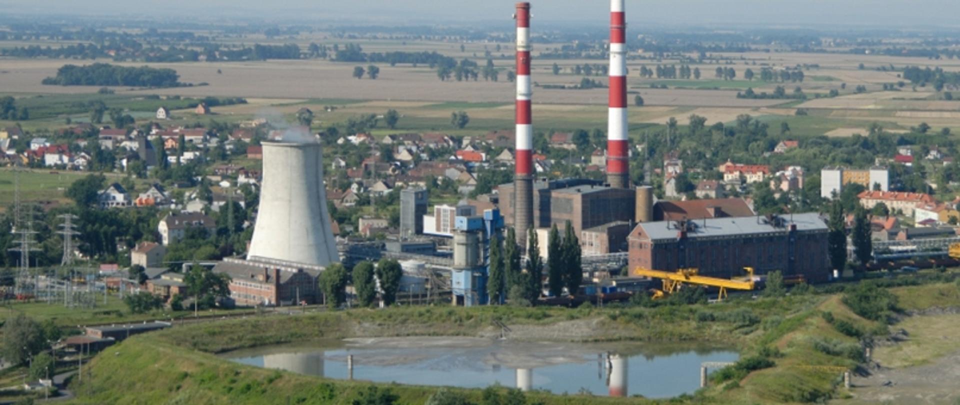 Widok na elektrociepłownię, kominy, budynki, zbiornik wodny z lotu ptaka. W pobliżu znajdują się pola i budynki mieszkalne.