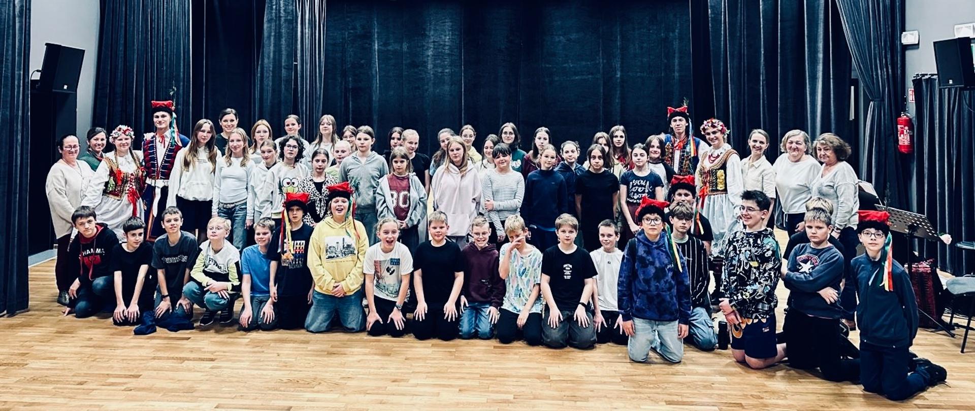 Na zdjęciu widać dużą grupę osób — dzieci, młodzież i dorosłych — ustawionych wspólnie do zdjęcia grupowego w sali teatralnej. W tle znajdują się ciemne zasłony sceniczne i oświetlenie teatralne skierowane na scenę.
Osoby stoją w kilku rzędach: z przodu siedzą lub klęczą młodsze dzieci, a z tyłu stoją starsze dzieci, młodzież i dorośli. Część osób ma na sobie kolorowe stroje śląskie i nakrycia głowy. Podłoga jest drewniana, a całość ma charakter pamiątkowego zdjęcia grupowego.