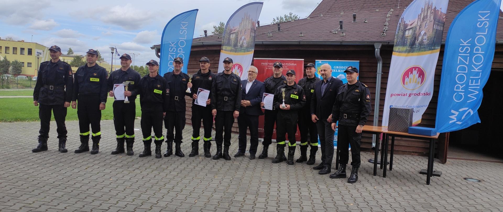 Na zdjęciu widać strażaka – kpt Andrzeja Śliwkę, który otrzymuje puchar przechodni mistrza wielkopolski strażaków PSP w strzelectwie sportowym. Puchar wręczany jest z rąk komendanta powiatowego PSP w Grodzisku Wielkopolskim. W tle widać pododdział strażaków podczas ceremonii wręczenia dyplomów oraz pucharów. 