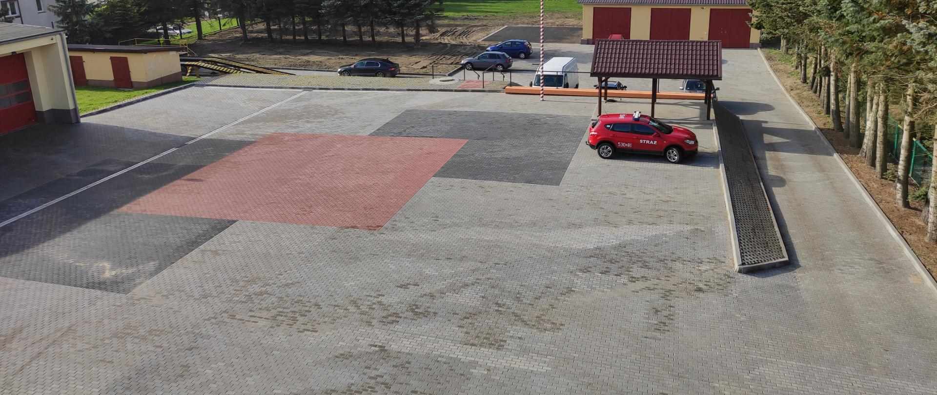 Plac manewrowy pokryty kostką brukową, znajduje się na nim samochód strażacki osobowy koloru czerwonego, masz na flagę w biało czerwone pasy oraz wiata na zabytkową bryczkę, na drugim planie samochody funkcjonariuszy