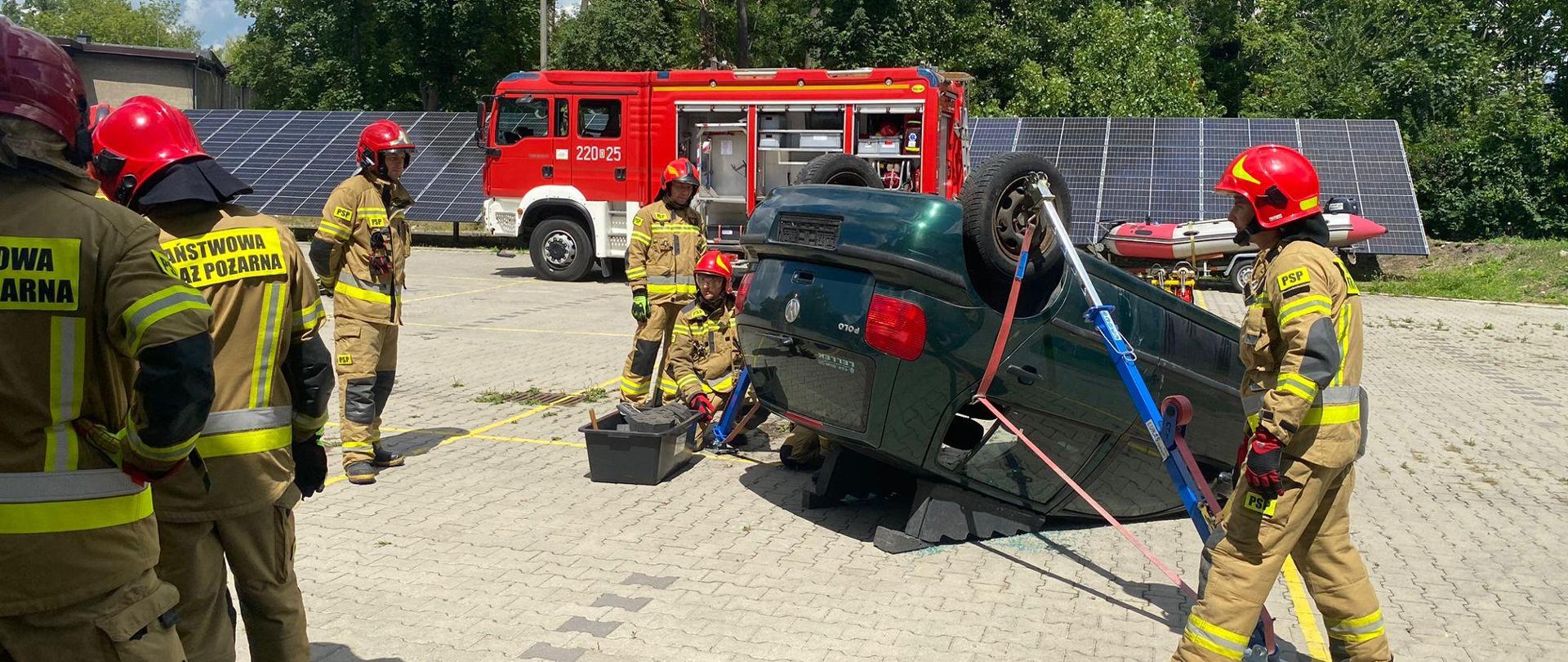 Strażacy Państwowej Straży Pożarnej w ubraniach bojowych i czerwonych hełmach na głowie ćwiczą na placu Komendy Wojewódzkiej Państwowej Straży Pożarnej założenia związane z ratownictwem technicznym. Strażacy przy użyciu różnych technik ratowniczych ćwiczą wykonanie dostępu do różnych części samochodu osobowego. W tym przypadku pojazd leżący na dachu.
