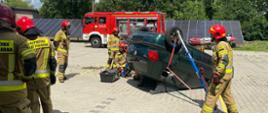 Strażacy Państwowej Straży Pożarnej w ubraniach bojowych i czerwonych hełmach na głowie ćwiczą na placu Komendy Wojewódzkiej Państwowej Straży Pożarnej założenia związane z ratownictwem technicznym. Strażacy przy użyciu różnych technik ratowniczych ćwiczą wykonanie dostępu do różnych części samochodu osobowego. W tym przypadku pojazd leżący na dachu.