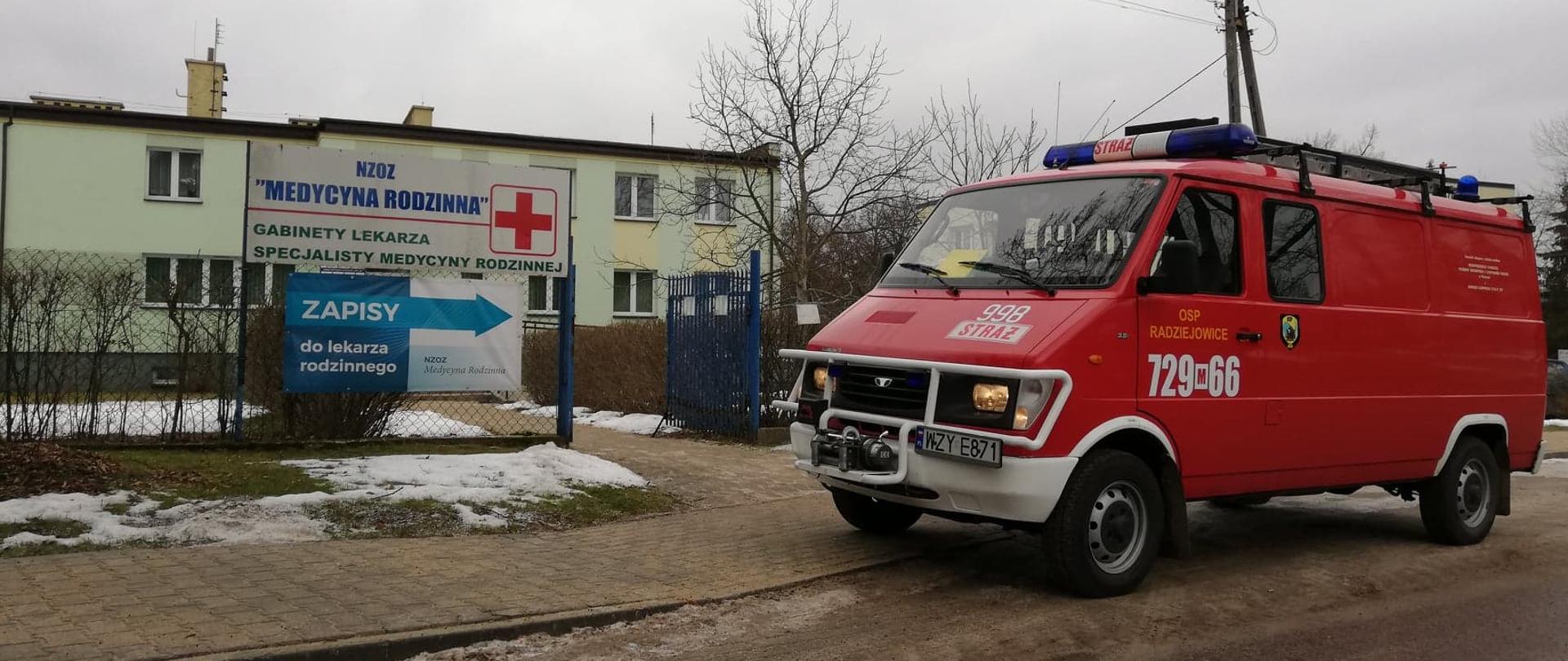 Na pierwszym planie stoi zaparkowany przy drodze czerwony samochód - strażacki bus z OSP Radziejowice. W tle widać blado zielony budynek przychodni.
