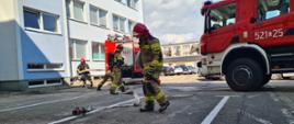 zdjęcie przedstawia 2 samochody strażackie przed budynkiem Urzędu Miejskiego w Sępólnie Krajeńskim. Pomiędzy autami znajduje się 4 strażaków w umundurowaniu specjalnym w trakcie rozwijania węży pożarniczych