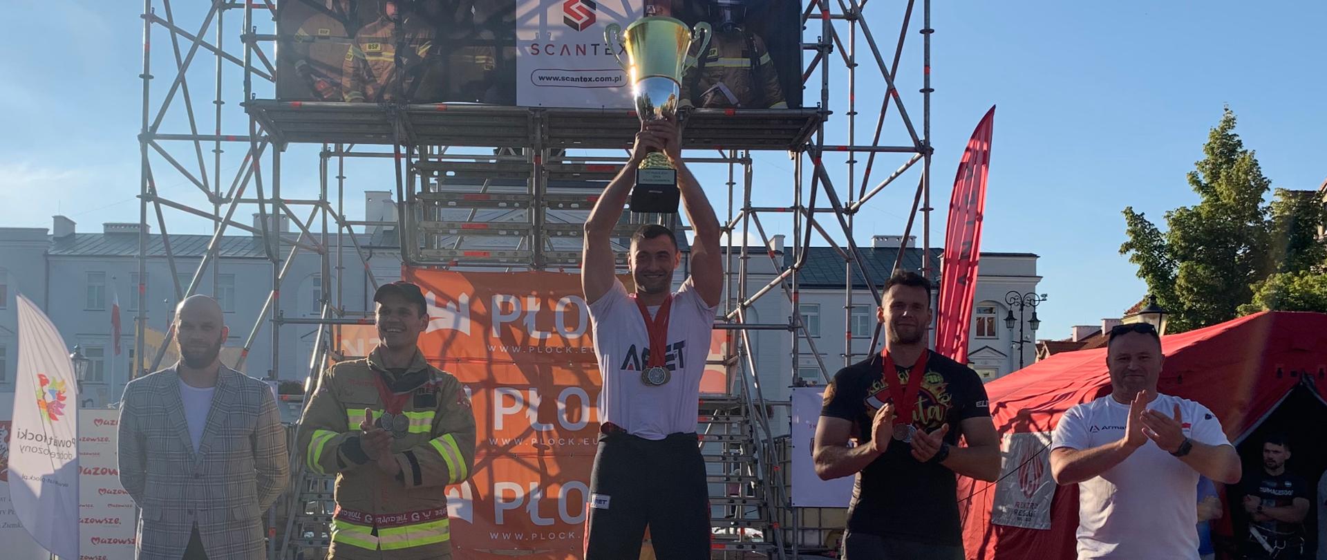 Adam na najwyższym stopniu podium wznosi puchar. Obok na innych miejscach stoi dwóch strażaków, a obok nich organizatorzy zawodów. W tle wieża TFC.