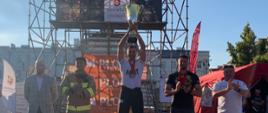 Adam na najwyższym stopniu podium wznosi puchar. Obok na innych miejscach stoi dwóch strażaków, a obok nich organizatorzy zawodów. W tle wieża TFC.
