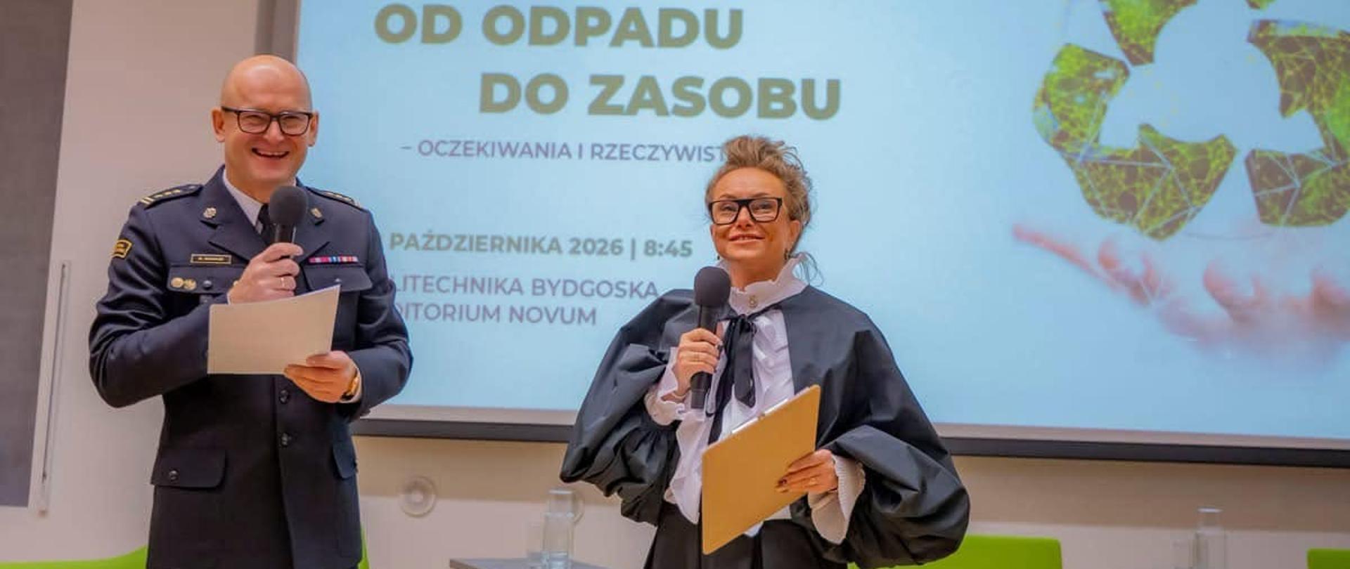 Kobieta i mężczyzna w mundurze prowadzą konferencję stojąc na scenie przed dużym ekranem.
