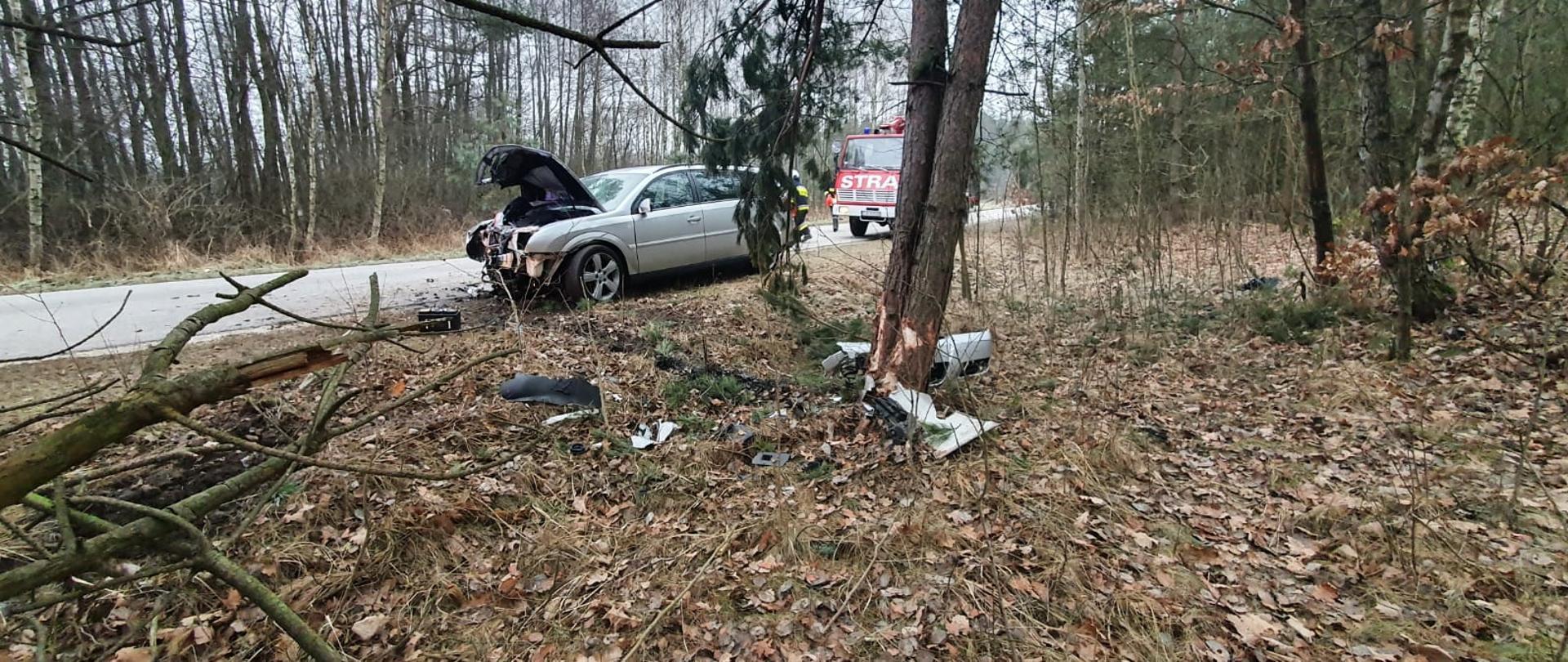 Zdjęcie przedstawia rozbity samochód osobowy na poboczu drogi powiatowej. w tle widnieje zaparkowany samochód ratowniczo-gaśniczy. Koło jednego z drzew widać elementy karoserii rozbitego pojazdu.