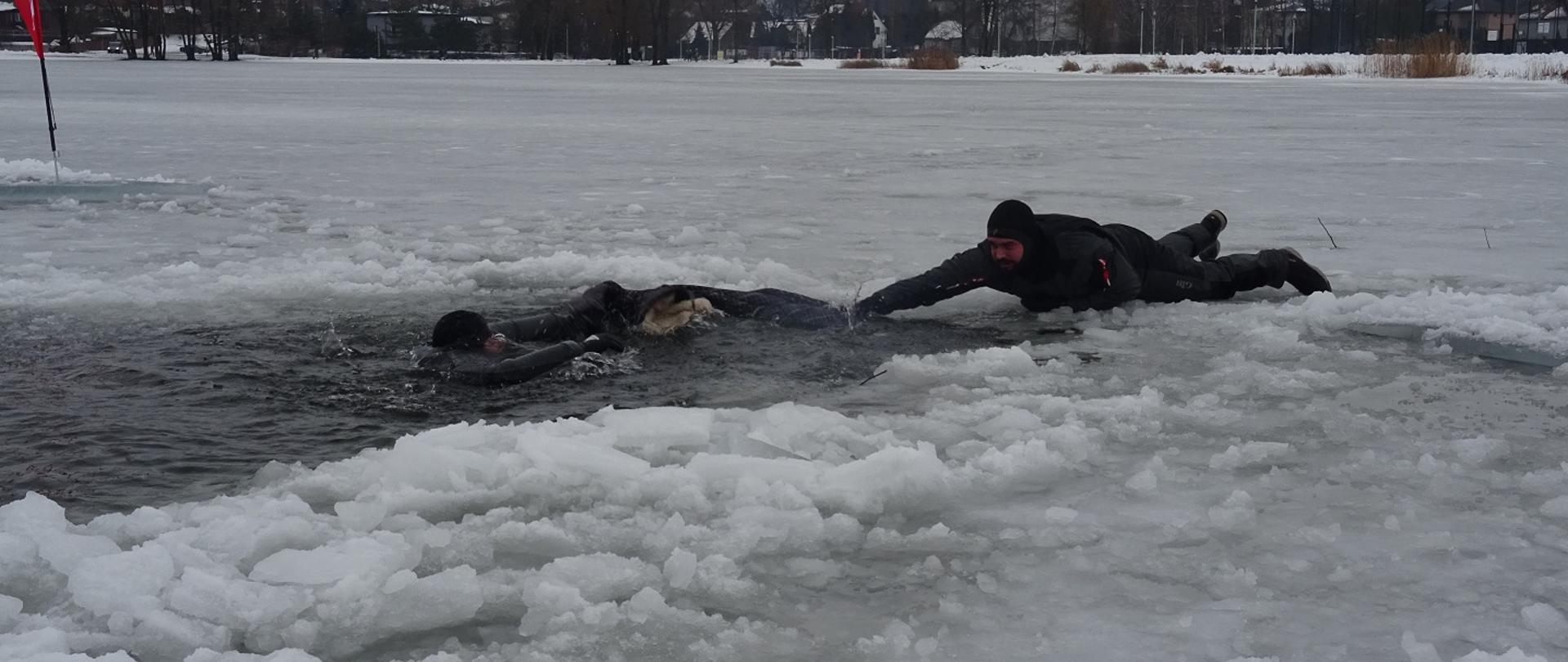 Zdjęcie obrazuje jedna osobę w przerębli i drugą która ja ratuje z powierzchni lodu