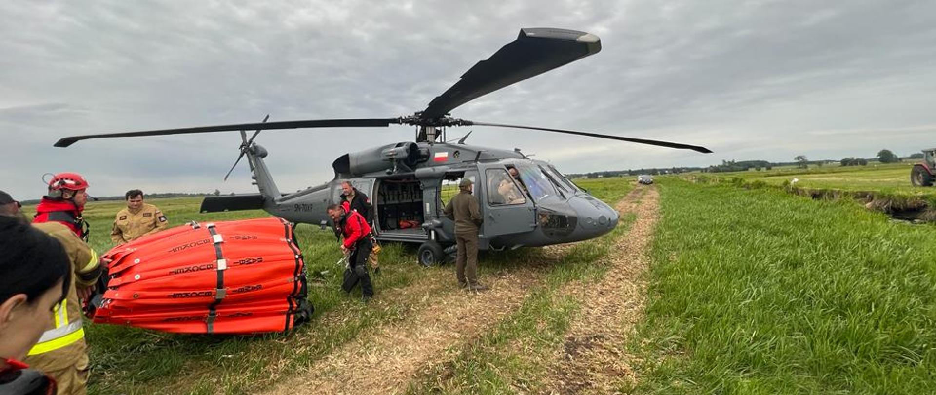 Na polanie stoi policyjny śmigłowiec z doczepionym z boku zbiornikiem na wodę typu Bambi Bucket oraz policyjni piloci i strażacy