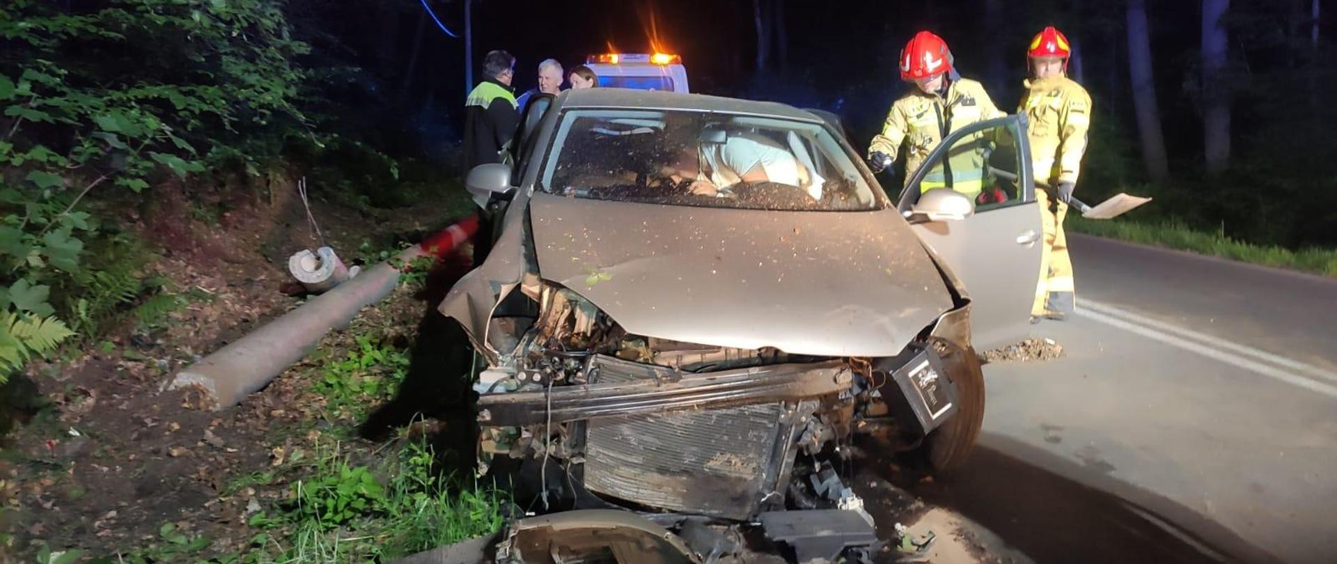 Samochód osobowy rozbity w wyniku wypadku drogowego, w tle strażacy oraz pozostałe służby oraz złamany słup elektryczny