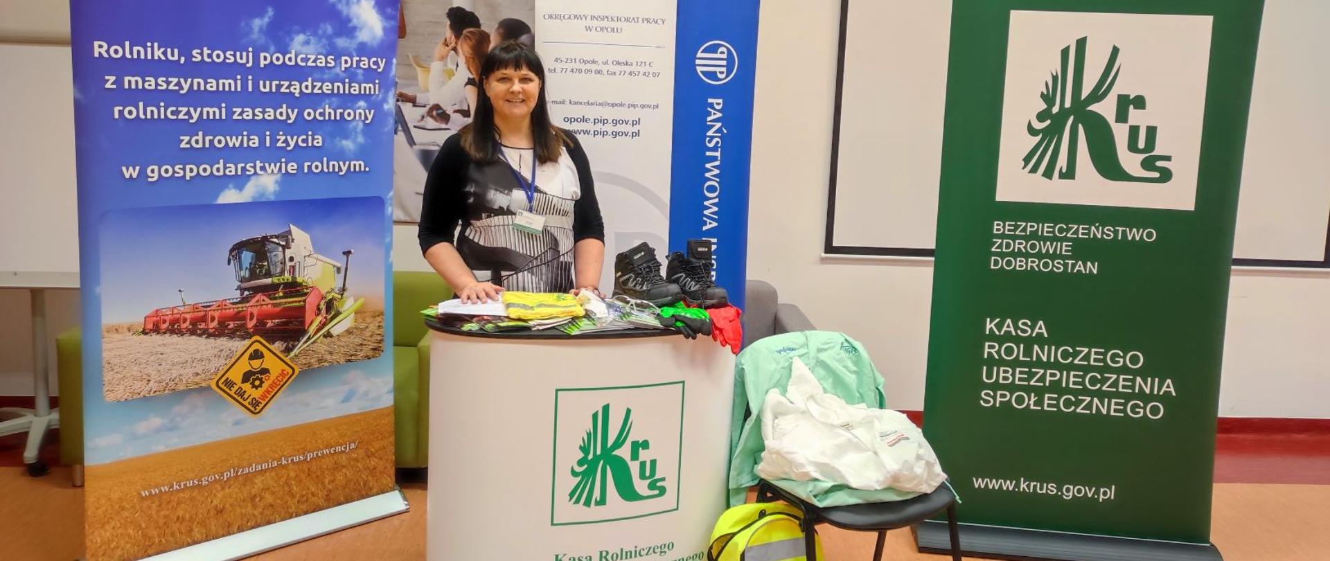 Młoda kobieta stoi za pulpitem z logo KRUS. Obok leżą elementy odzieży ochronnej, plecak odblaskowy. z tyłu stoją rollupy z hasłami kampanii KRUS i z logo KRUS.