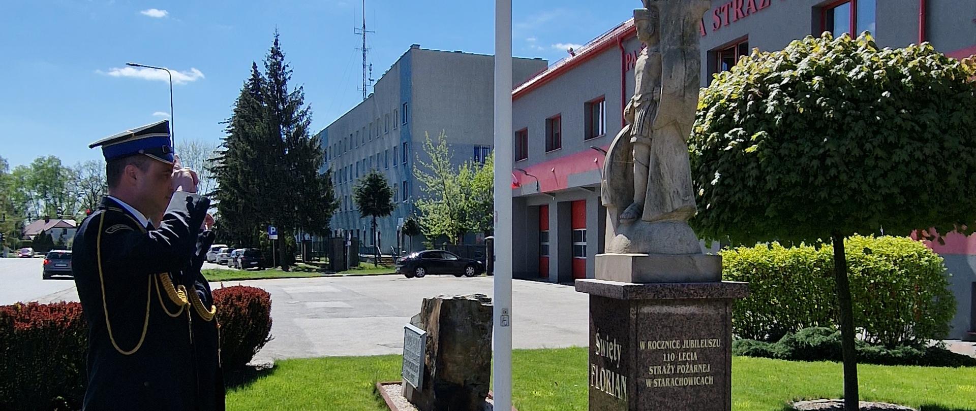 Strażacy oddają honor pod posągiem św. Floriana w tle budynek KP PSP