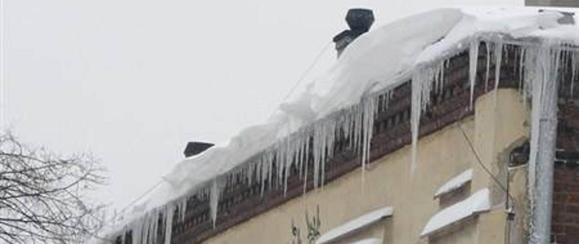 Na zdjęciu fragment dużego budynku, na którego dachu leży gruba warstwa śniegu, a z dachu zwisają duże sople lodu.