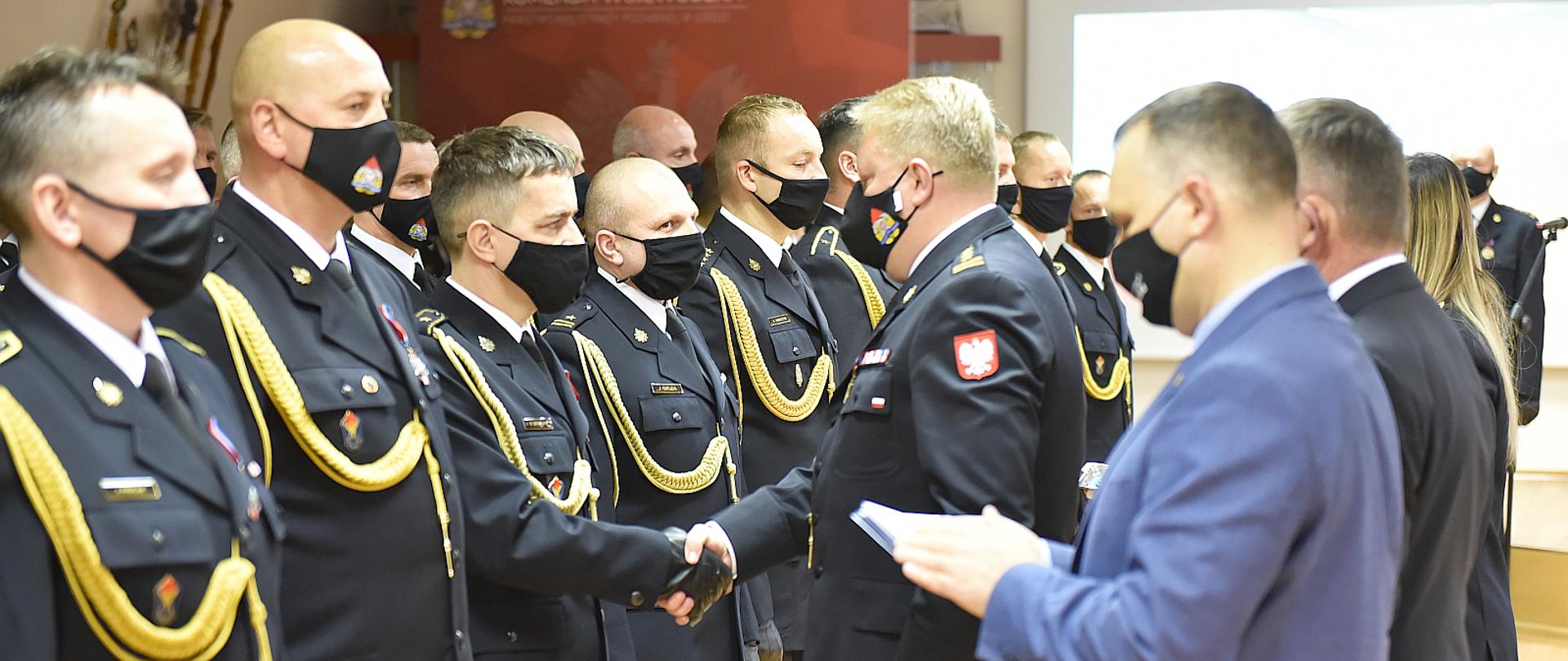 na zdjęciu grupa strażaków w umundurowaniu galowym stojąca na zbiórce w sali, jeden strażak stoi przez resztą strażaków i składa gratulacje poprzez uścisk dłoni, na zdjęciu widać również dwóch mężczyzn w garniturach są rozmazani, w tle widać drzewce sztandarów i baner komendy wojewódzkiej psp w łodzi