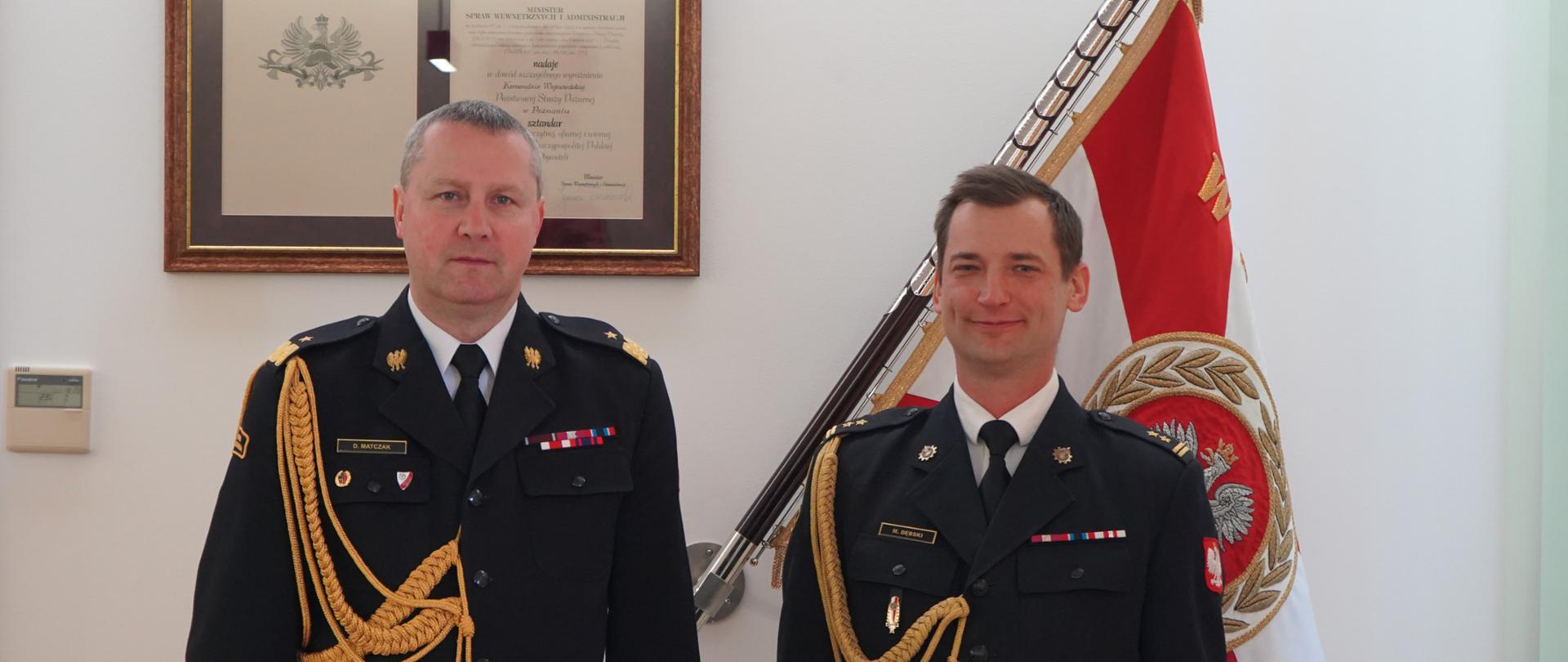 dwóch strażaków w ciemnych mundurach ze sznurem stoi obok siebie, w tle sztandar