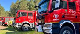 Pojazdy gaśnicze straży pożarnej ustawione na leśnym parkingu. W tle widać strażaków w ubraniach specjalnych.