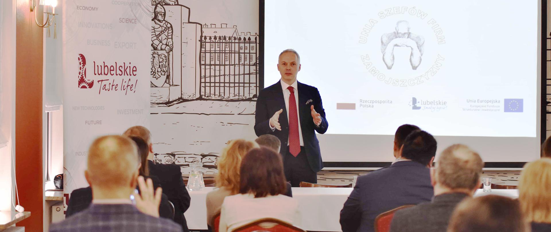 Wiceminister Jacek Żalek stoi przodem do siedzących na krzesłach ludziach. Za nim duży ekran.