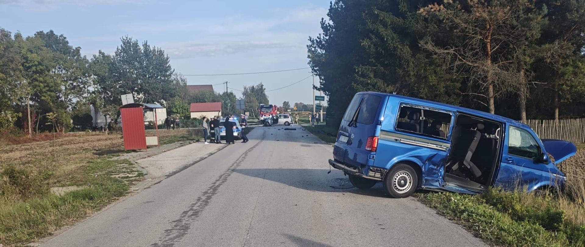 Przedstawia szeroki plan drogi objętej działaniami służb ratunkowych. Na pierwszym planie na asfalcie stoi żółty, trójkątny znak ostrzegawczy z napisem „WYPADEK” oraz pomarańczowe pachołki drogowe wyznaczające strefę zagrożenia. Po prawej stronie w rowie stoi wspomniany wcześniej niebieski van, a w głębi drogi widać radiowozy policyjne i inne pojazdy służb. 