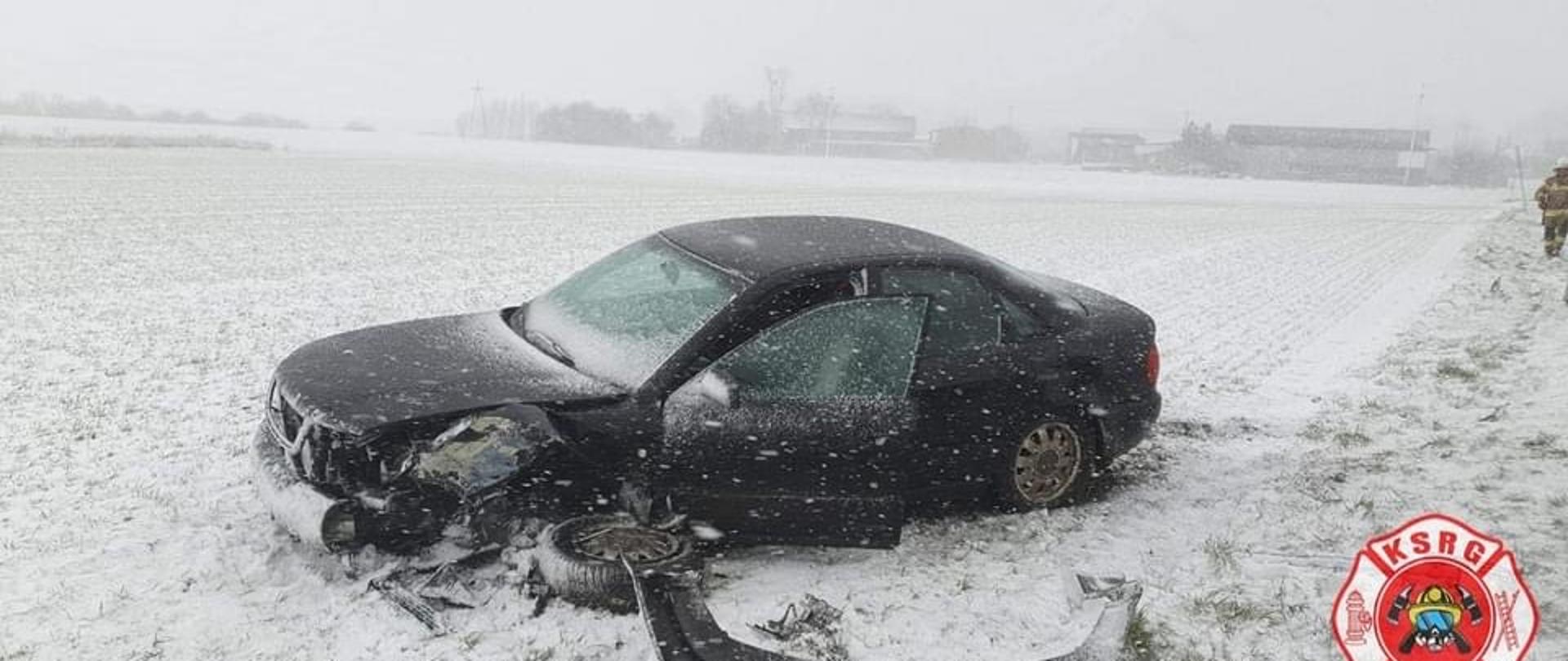 Samochód osobowy po kolizji na zaśnieżonym polu. Samochód ma oderwane lewe koło i zderzak