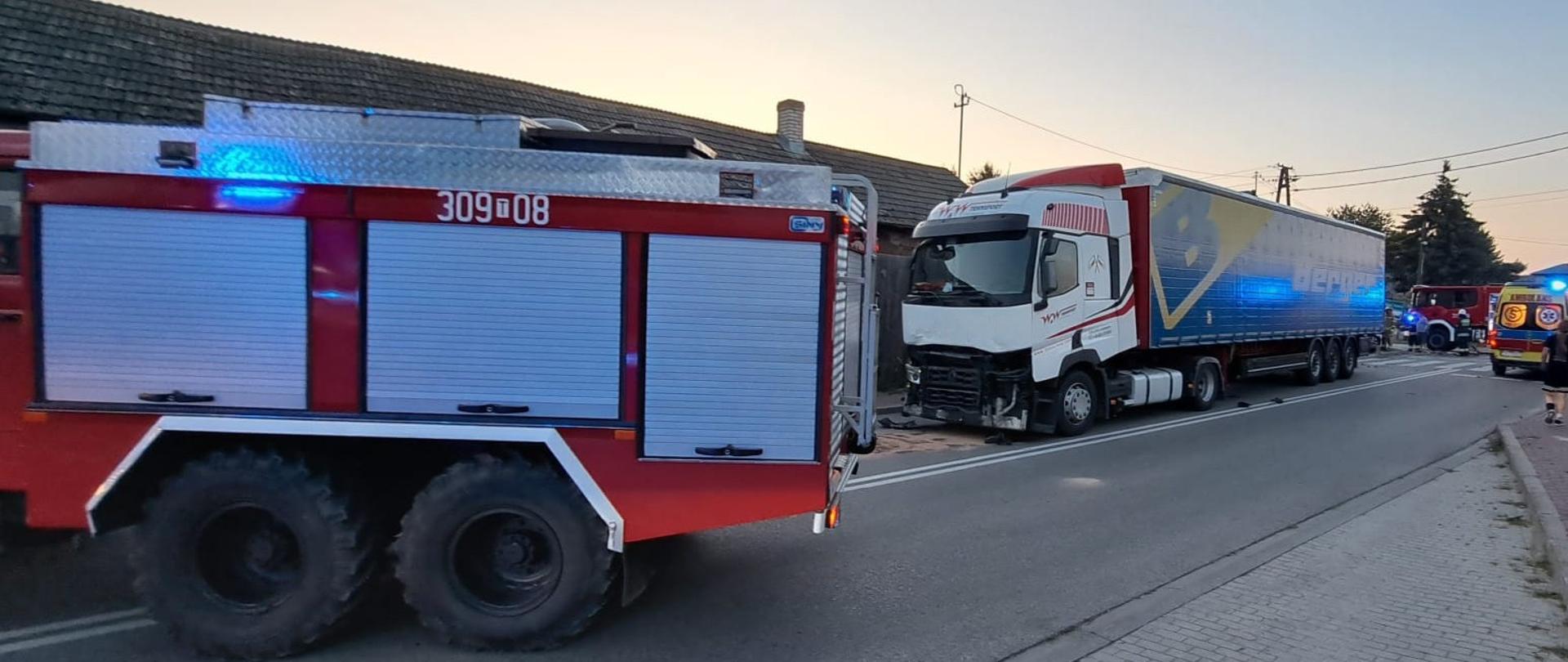 Zdjęcie przedstawia miejsce wypadku: z lewej strony samochód pożarniczy, dalej uszkodzony samochód ciężarowy, dalej drugi pojazd strażaków i po prawej ambulans.