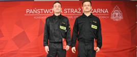 Dwóch strażaków w czarnych mundurach stoi na tle czerwonej ścianki z napisem ‘Państwowa Straż Pożarna – Komenda Miejska Państwowej Straży Pożarnej w Żorach’ oraz logo PSP. Patrzą prosto w kamerę i trzymają dokumenty w prawej ręce. 