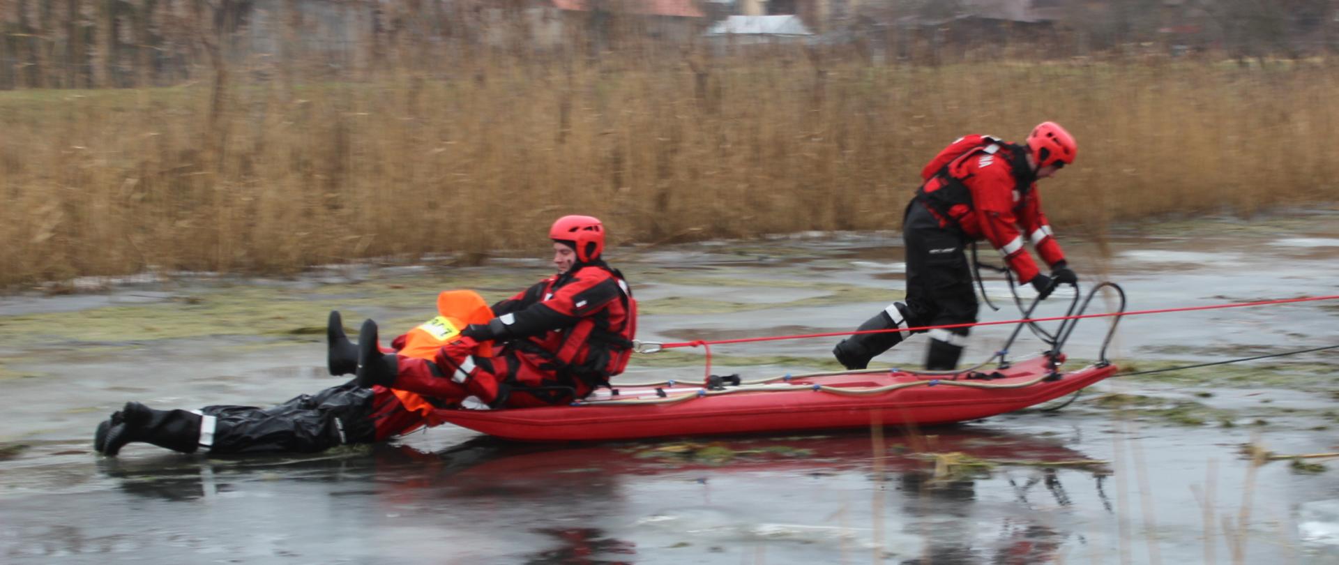 Dwóch strażaków podczas pokazów ratownictwa wodno-lodowego prezentuje metodę wydobycia poszkodowanego przy użyciu sań lodowych. Strażacy oraz pozorant ubrani są w specjalistyczne skafandry do ratownictwa wodnego.