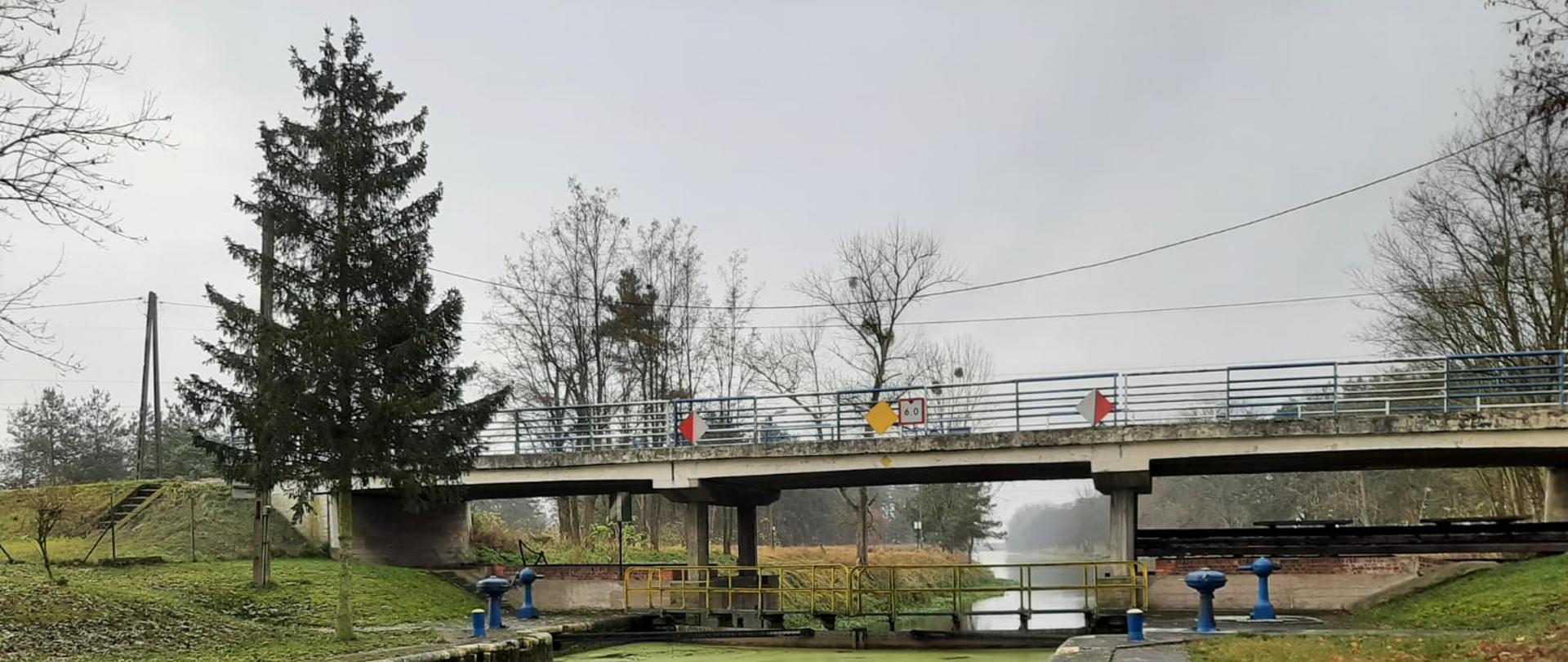 Śluza Gromadno - ostatnia dziesiąta śluza w województwie kujawsko-pomorskim. Na pierwszym planie komora śluzy napełniona wodą, na drugim planie zamknięte wrota śluzy, nad wrotami most drogowy, w oddali mgła spowijająca rzekę Noteć. Jesień. Po obu stronach nabrzeża leżą żółto-brązowe liście. 