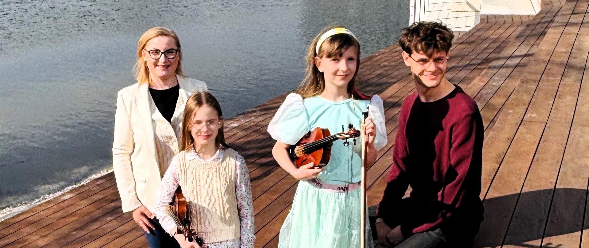 Cztery osoby pozujące (dwie z nich trzymają skrzypce) na drewnianym tarasie nad wodą, na tle nowoczesnego, przeszklonego budynku w słoneczny dzień.