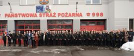 Strażacy oraz zaproszeni goście pozują do fotografii stojąc przed budynkiem komendy.
