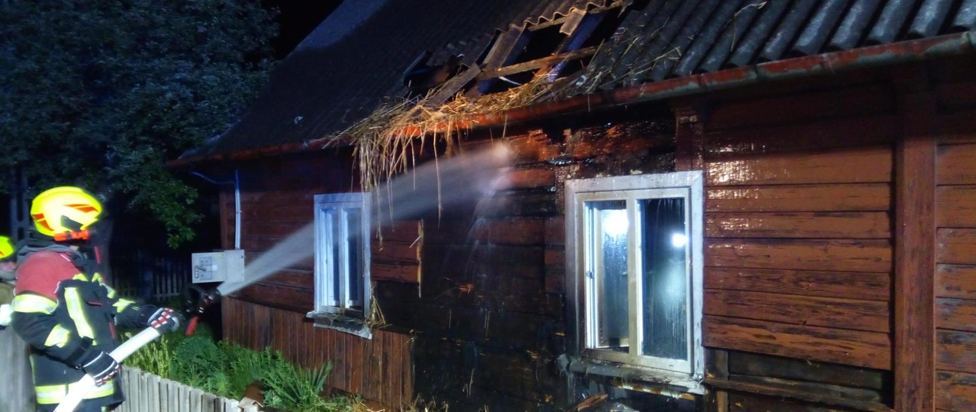 Zdjęcie przedstawia strażaka podającego wodę na palącą się ścianę budynku jednorodzinnego.