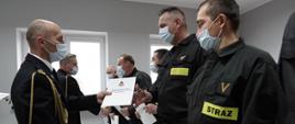 Komendant powiatowy mł. bryg. Arkadiusz Makowski ubrany w mundur galowy ze sznurem w maseczce ochronnej na twarzy wręcza awanse strażakom tutejszej komendy, w lewej dłoni trzyma teczkę na akta, w głębi Zastępca komendanta powiatowego w Łowiczu mł. bryg. Rafał Garbarz ubrany w mundur galowy ze sznurem w maseczce ochronnej na twarzy składa gratulacje funkcjonariuszom, strażacy stoją w szeregu w pozycji zasadniczej ubrani w ubranie koszarowe w maseczkach ochronnych na twarzy