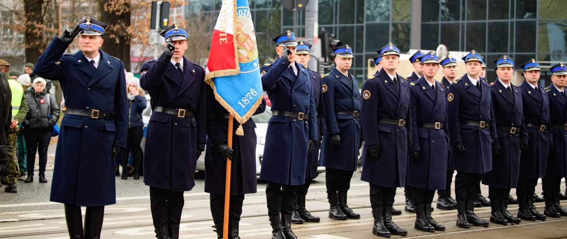 Szereg umundurowanych funkcjonariuszy w długich, granatowych płaszczach, ustawionych na zewnątrz, wszyscy salutują. Pośrodku oficer trzyma sztandar w kolorach czerwonym, białym i niebieskim.