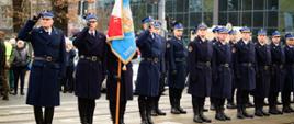 Szereg umundurowanych funkcjonariuszy w długich, granatowych płaszczach, ustawionych na zewnątrz, wszyscy salutują. Pośrodku oficer trzyma sztandar w kolorach czerwonym, białym i niebieskim.