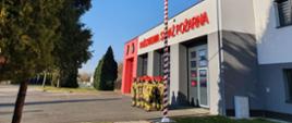 Strażacy w mundurach bojowych stoją w dwuszeregu na tle szarej ściany komendy. Po ich lewej stronie powiewa wciągnięta na maszt flaga państwowa. Po lewej stronie zdjęcia drzewo iglaste.
