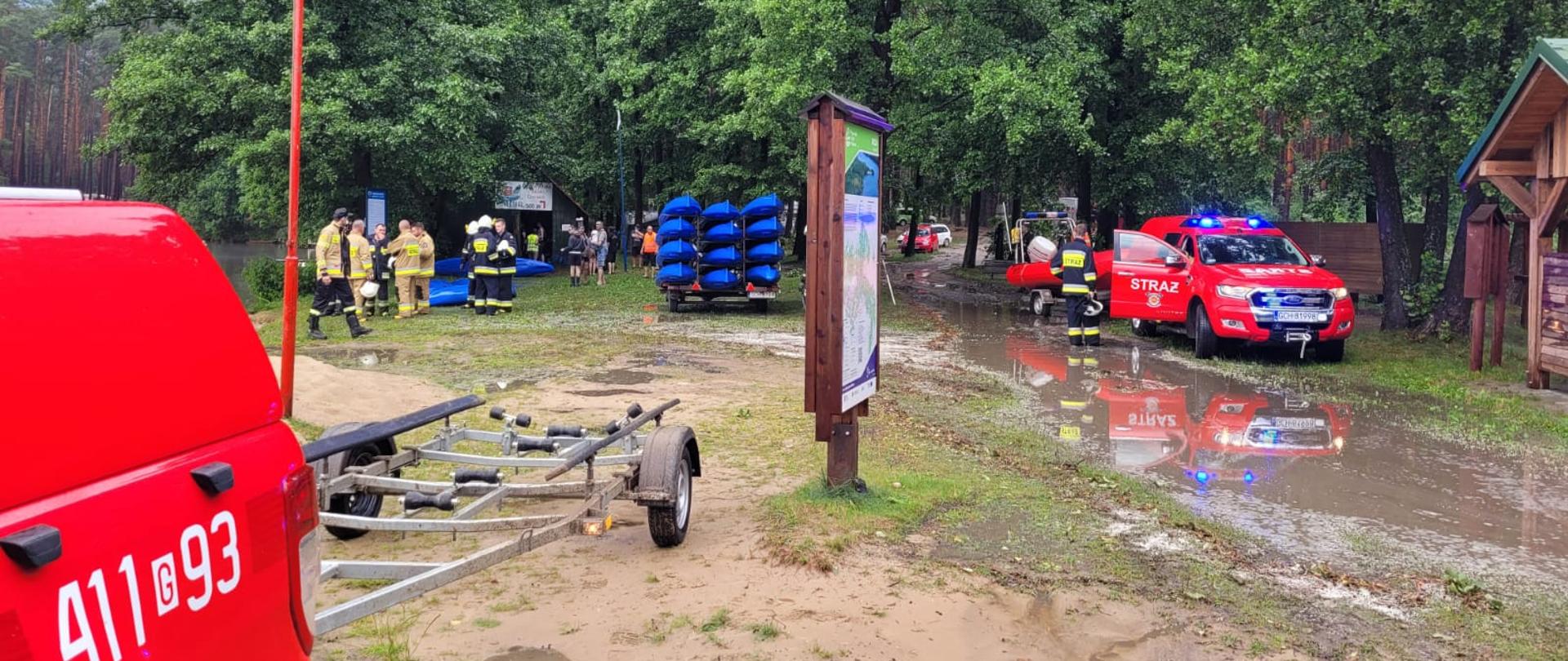 Brzeg jeziora, kałuże po ulewie, widoczne wozy strażackie i przyczepka do przewozu łodzi. W tle zdjęcia widocznej kajaki. W oddali pracujący strażacy. 